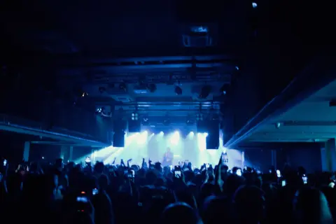 Tłum na koncercie lil tracy, ludzie nagrywają telefonami, światła sceniczne tworzą niebieską poświatę.