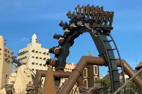 A roller coaster with brown seats and patterned spheres against a blue sky and themed buildings.