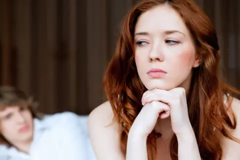 Traurige Frau mit roten Haaren blickt weg. Hinter ihr liegt ein Mann im Bett. Sie spürt keine sexuelle Anziehung mehr zum Partner.