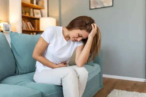 Femme assise sur un canapé bleu, se tenant le ventre et la tête, visiblement souffrante. Elle pourrait exprimer la douleur liée à l'endométriose profonde.
