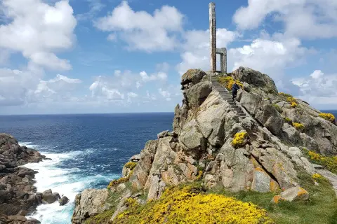 Escaleras de piedra ascienden por rocas cubiertas de flores amarillas hasta un monumento en el cabo Priorino. El mar azul y el cielo con nubes completan la escena.
