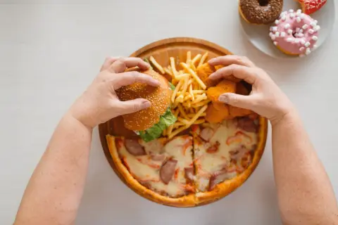 Mani che afferrano un hamburger, patatine, nuggets e pizza. Un vero **cheat day** con ciambelle in lontananza.
