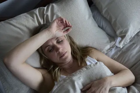 Mujer de unos 40 años sufre sudores nocturnos, con la mano en la frente, en la cama.