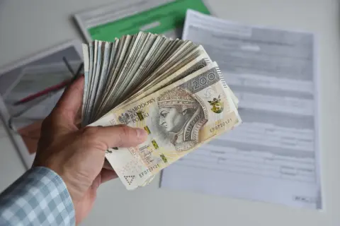 Dłoń trzyma plik banknotów 200 zł, symbolizujących zarobki, być może nawet najniższą krajową netto, na tle dokumentów.