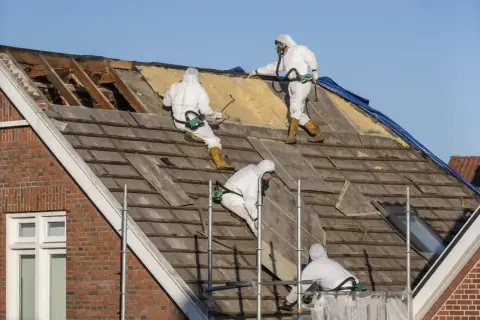 Pracownicy w kombinezonach usuwają stare pokrycie dachu, które może zawierać azbest. Co to jest azbest i dlaczego jest niebezpieczny?