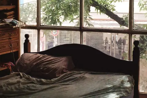 Dormitorio con ventana en el cabecero. La cama está hecha, con sábanas arrugadas y una almohada. A la izquierda, un mueble de madera con cajones.