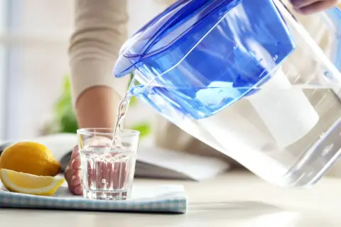 Une personne verse de l'eau filtrée d'une carafe bleue dans un verre. À côté, un citron coupé. L'eau filtrée est elle bonne pour la santé ?