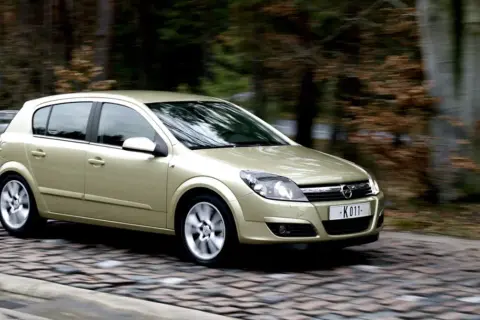 Złoty Opel Astra H 1.7 CDTI w ruchu na brukowanej drodze, otoczony rozmazanym tłem lasu.