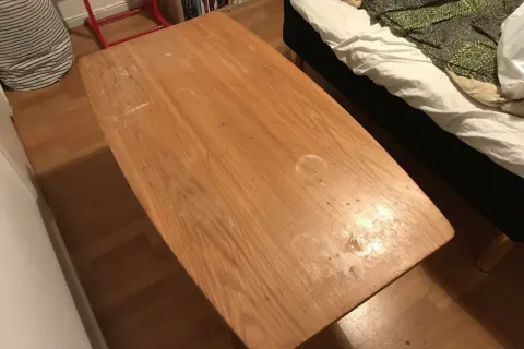 Ett slitet soffbord i trä med fläckar, redo att måla om. Bredvid syns en säng med vitt och grönt överkast.