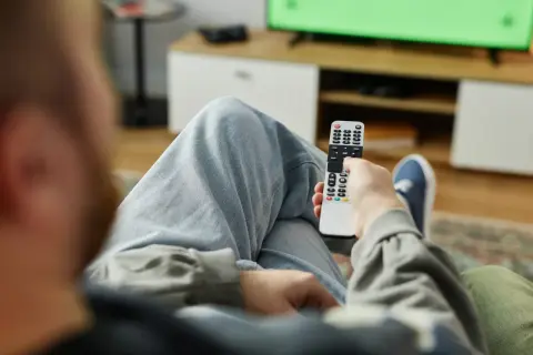 Mężczyzna z pilotem w ręku, zastanawia się, kto nie musi płacić abonamentu RTV, oglądając zielony ekran telewizora.