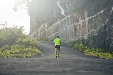 Un coureur en tenue fluo grimpe une route sinueuse, affrontant un dénivelé positif de 100 m, bordée de végétation luxuriante et d'une falaise imposante.
