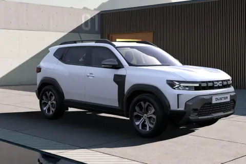 Nowa Dacia Duster w białym kolorze prezentuje się nowocześnie na tle betonowej ściany i budynku z drewnianymi elementami.