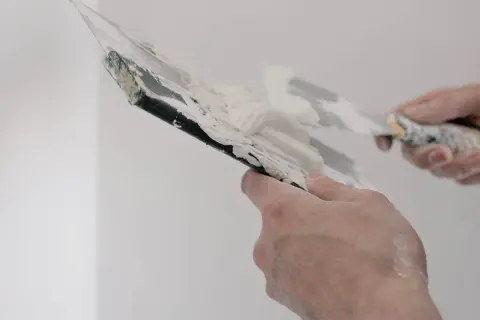 Händer som spacklar en vägg. Spacklet är vitt och klibbigt, och verktygen är täckta av det.