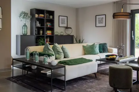 Gemütliches Wohnzimmer mit cremefarbener Couch, grünen Kissen und Decken. Helle Farben dominieren, ergänzt durch dunkle Möbel und eine große Glasfront.