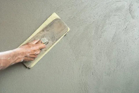 Mano che stende l'intonaco con una spatola, creando una finitura liscia. La rasatura 5 mm è quasi completa.