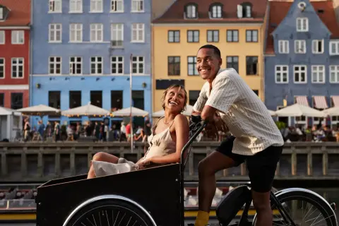 Ett lyckligt par njuter av en romantisk weekend i Europa, cyklar längs en kanal med färgglada hus i bakgrunden.