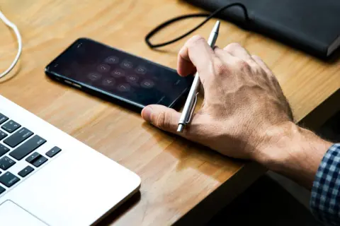 Dłoń trzyma długopis nad ekranem z klawiaturą numeryczną, gdzie urządzenie iPhone niedostępne. Obok leży laptop.