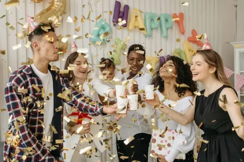 Fröhliche Freunde feiern eine Geburtstagsparty mit Konfetti und Luftballons. Ein "Happy Birthday"-Banner hängt im Hintergrund. Ein tolles Lied für Erwachsene!