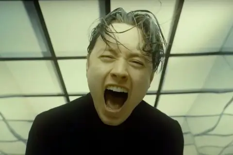 Noah Sebastian screams in a black shirt, his hair wet and messy, against a backdrop of geometric patterns.