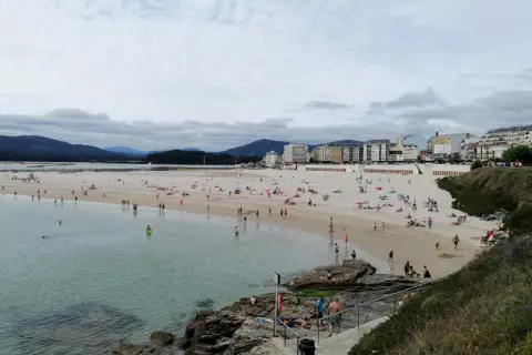 Gente disfruta de un día soleado en la playa de peizas, con aguas cristalinas y edificios al fondo.