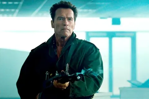 Arnold Schwarzenegger w zielonej kurtce z karabinem, gotowy do akcji w jednym ze swoich kultowych filmów.