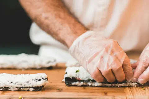 Hände mit Handschuhen rollen Sushi. Reis, Nori und Gemüse sind die Zutaten für sushi selber machen.