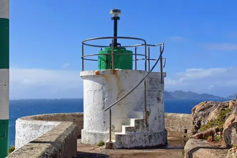 Faro blanco con base verde y escaleras externas, junto al mar azul. El **monte Ferro** se divisa al fondo.