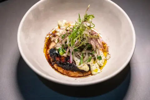 Plato gourmet con un trozo de carne oscuro y crujiente, cubierto de cebolla morada y hierbas frescas, bañado en una salsa dorada. Un solete Repsol para el paladar.