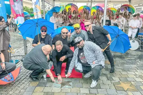 Grupa mężczyzn, prawdopodobnie zespół, odsłania gwiazdę z napisem "PIERSI BAŁKANICA" na chodniku. W tle kolorowe parasole i publiczność.