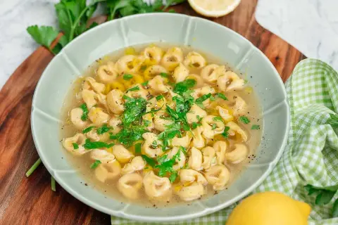 En skål med tortellini in brodo, garnerad med färsk persilja och en citronskiva.