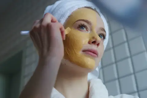Junge Frau mit Handtuch auf dem Kopf trägt eine Kurkuma-Maske auf. Vorher-Nachher-Effekt für strahlende Haut.