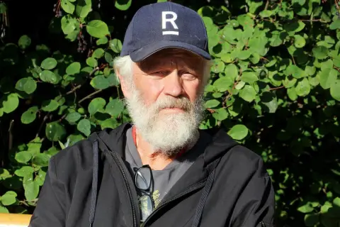 En äldre man med vitt skägg och keps med ett R på sitter utomhus. Han är en del av Ronny Eriksson familj.