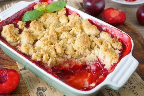 Ein warmer pflaumen crumble mit knuspriger Streuseldecke und frischer Minze. Daneben liegen saftige Pflaumen.