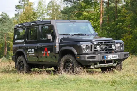 Czarny grenadier auto z bagażnikiem dachowym i snorkelami, zaparkowany na trawie wśród drzew.