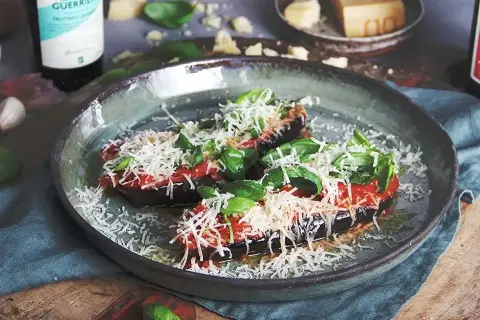 Två halvor av aubergine i ugn, toppade med tomatsås, basilika och riven ost. Serveras på en rustik tallrik.