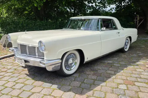 Ein cremefarbener amerikanischer Auto-Klassiker, ein Lincoln Continental, steht auf einem Kopfsteinpflasterweg unter Bäumen.