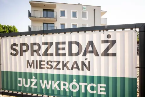 Baner z napisem "SPRZEDAŻ MIESZKAŃ JUŻ WKRÓTCE" na tle nowoczesnego budynku mieszkalnego.