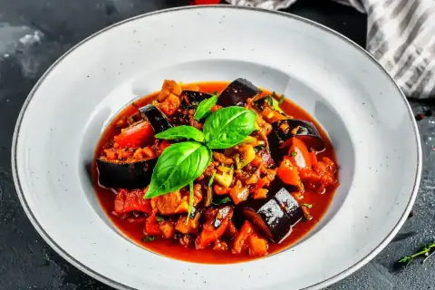 Un plat de ratatouille au four, garni de basilic frais, avec des aubergines, des tomates et d'autres légumes mijotés dans une sauce savoureuse.