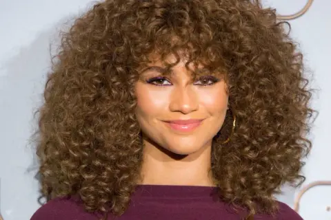 Mujer con cortes pelo rizado largo y voluminoso, maquillaje vibrante y sonrisa radiante.