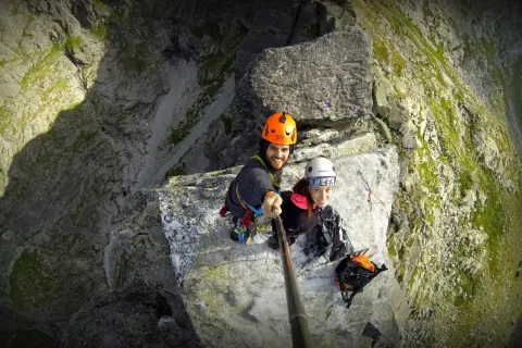 Uśmiechnięci alpiniści na skalnej półce, gotowi na wejście na mnicha. Widok z góry na stromą ścianę.