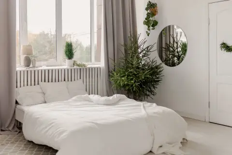 Biała sypialnia inspiracje: przytulne łóżko z pościelą, choinka, rośliny i lustro tworzą spokojną atmosferę.