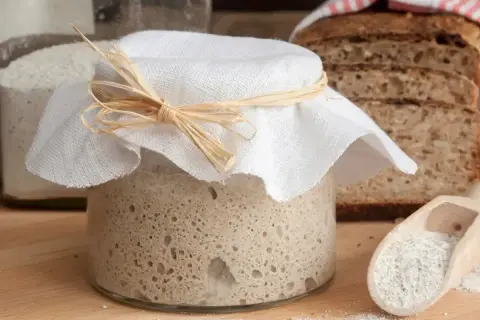 Słoik z zakwasem, gotowy do pieczenia chleba. Obok kromki pysznego, lekko lepkiego w środku chleba.