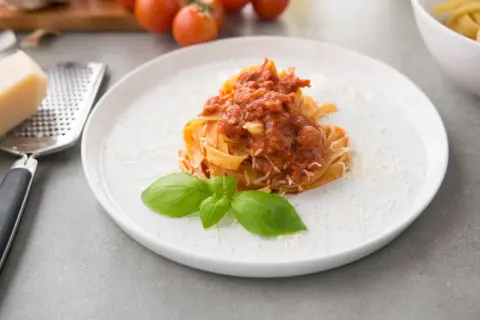 Talerz z makaronem tagliatelle polanym sosem pomidorowym, posypany parmezanem i udekorowany bazylią.