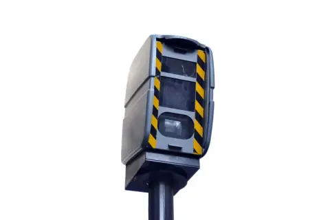 Boîtier gris avec bandes noires et jaunes, monté sur un poteau. Ce radar de contrôle de vitesse est un modèle différent de ceux que l'on voit habituellement.