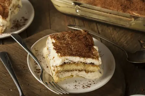 Une part de tiramisu, dessert dont l'origine est débattue, sur une assiette avec une fourchette. Le dessert est saupoudré de cacao.