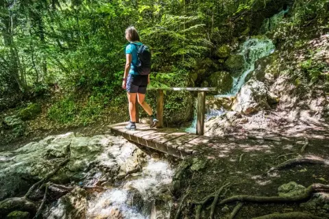 Une randonneuse traverse un pont en bois près de la cascade de Morette, entourée d'une végétation luxuriante.