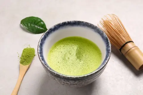 Bol rempli de matcha mousseux, une cuillère en bois avec de la poudre verte et un fouet en bambou. Une feuille de thé verte repose à côté, évoquant le pouvoir antioxydant du matcha.
