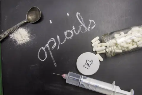 Napis "opiaty" na tablicy, obok łyżki z proszkiem, strzykawki i butelki z tabletkami.