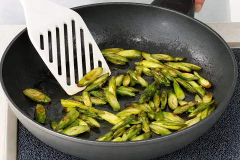 Geschnittener Spargel brät in einer Pfanne. Ein weißer Pfannenwender bewegt die grünen Stücke.