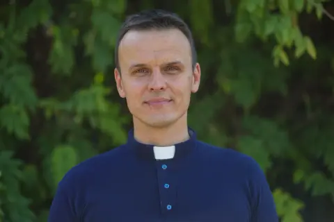 Młody ksiądz z osiedla w granatowej koszuli z kołnierzykiem, uśmiecha się na tle zieleni.